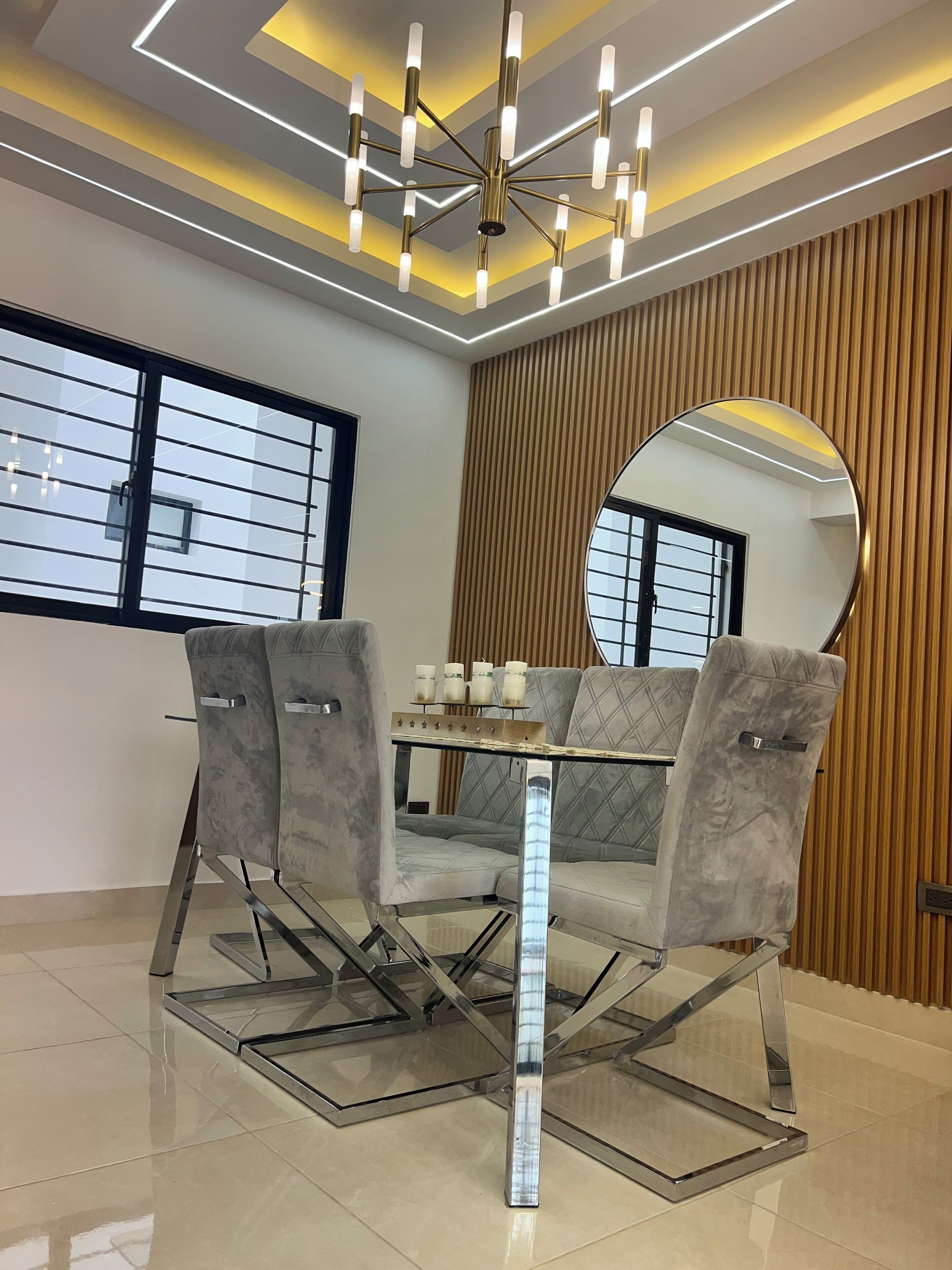 Un espace de salle à manger moderne avec une table en verre, des chaises à revêtement gris, un lustre central et un grand miroir mural.