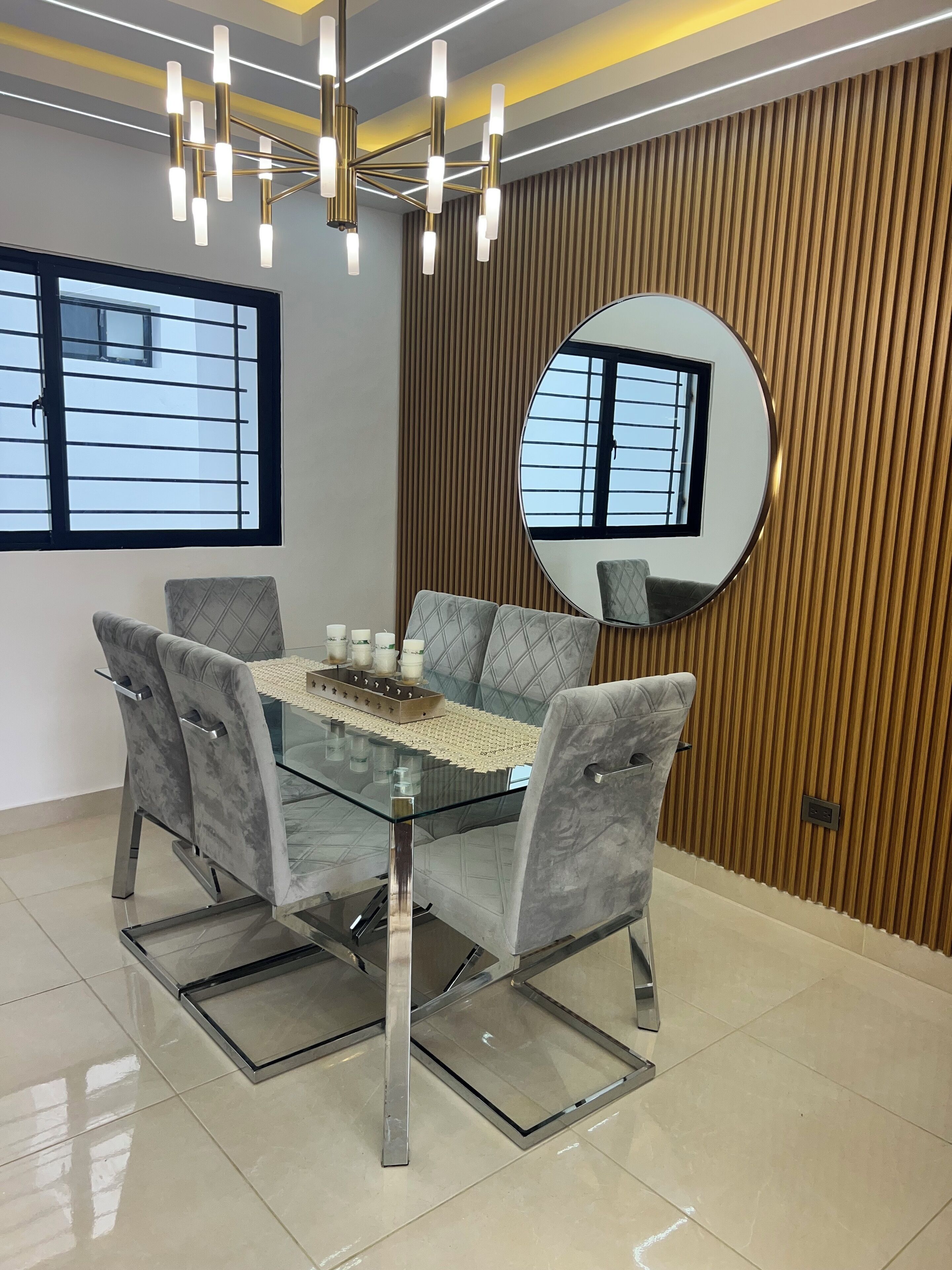 Een moderne eetruimte met een glazen tafel, grijs gestoffeerde stoelen en een decoratieve spiegel aan de muur.