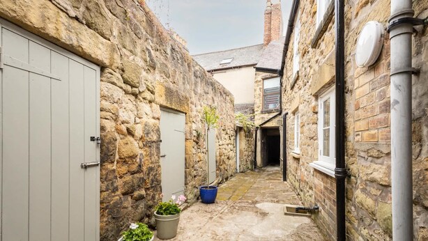 A narrow stone alleyway with a door on the left, a blue bucket, and two potted plants.