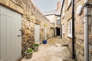 A narrow stone alleyway with a door on the left, a blue bucket, and two potted plants.