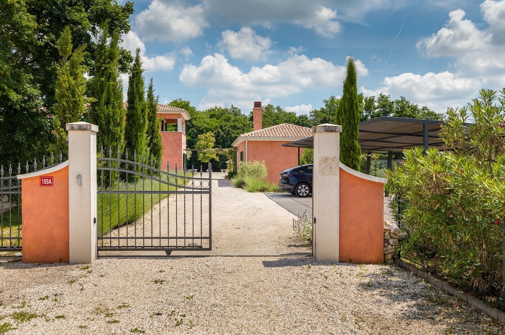 A gated entrance to a property with a gravel driveway, a parked car, and a building with a tiled roof.