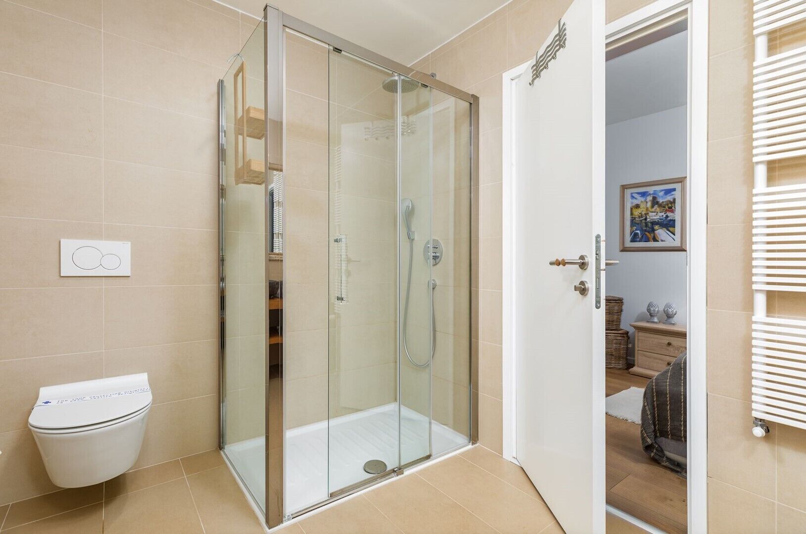 A modern bathroom with a glass-enclosed shower, a toilet, and a door leading to a bedroom with a bed and a dresser.
