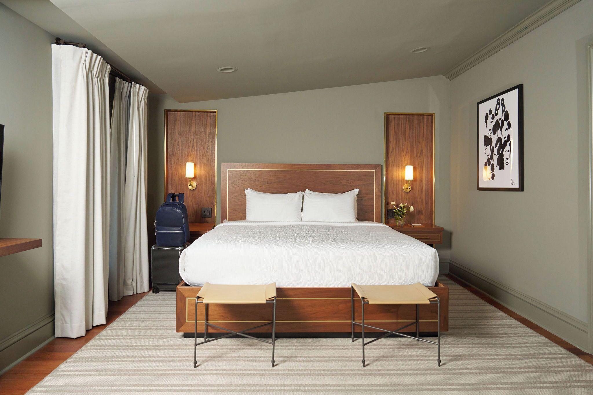 A hotel room with a large bed, bedside lamps, a wooden headboard, a bench, a suitcase, and a framed artwork on the wall.