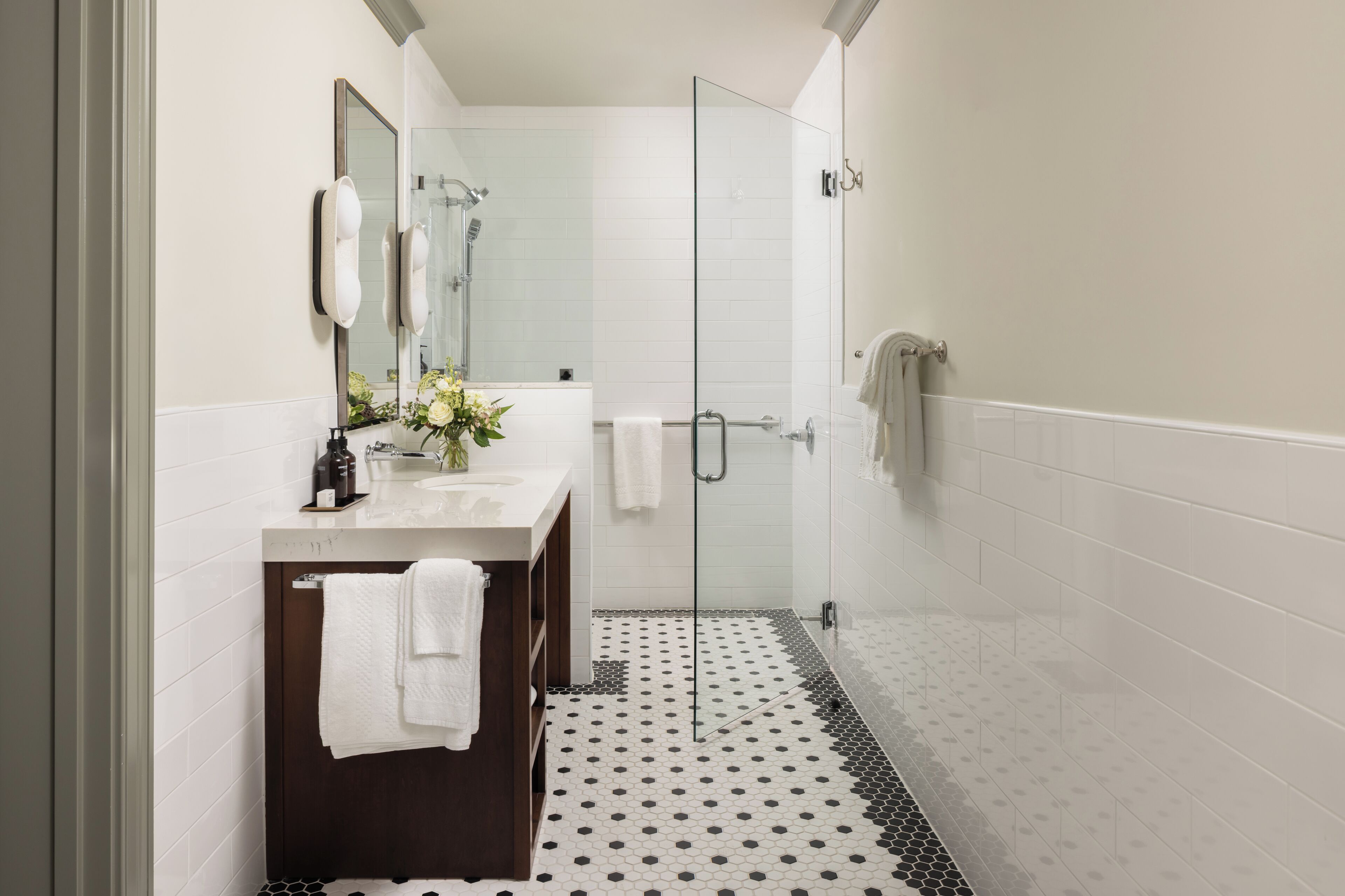 A modern bathroom with a glass shower, white tiles, a dark wooden vanity, and a black and white patterned floor.
