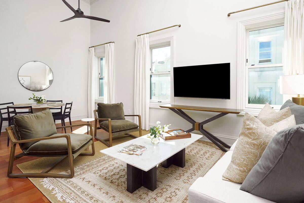 A modern living room with a flat-screen TV, two armchairs, a sofa, a coffee table, and a dining area with chairs and a table.
