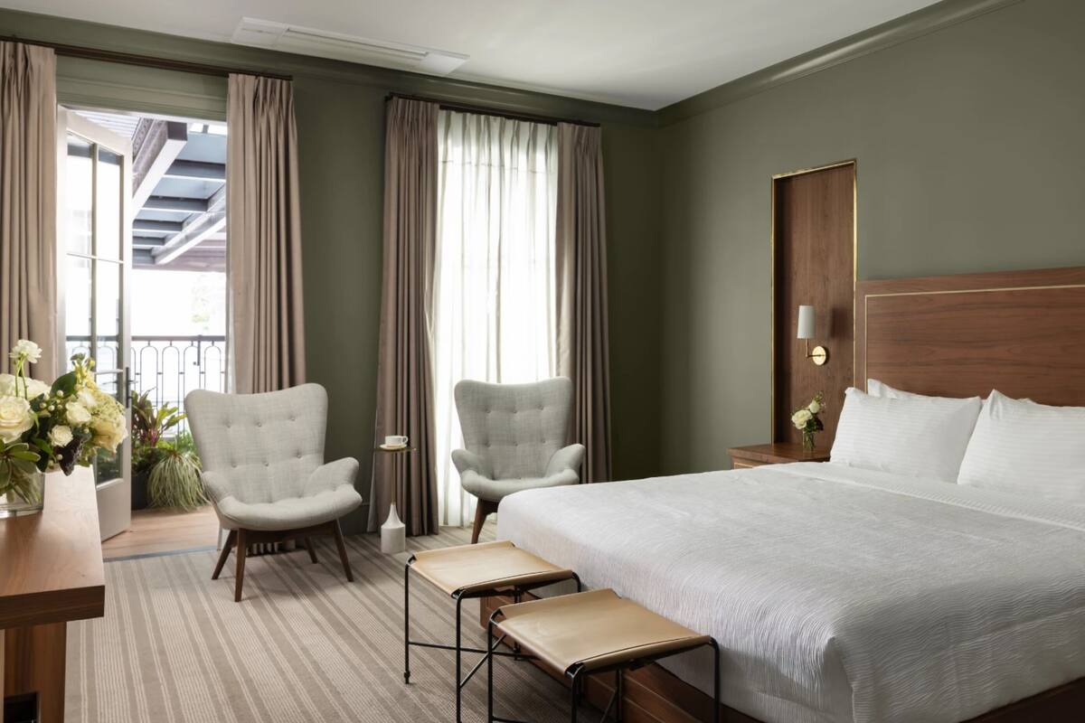 A hotel room with a large bed, two armchairs, a small table, and a nightstand with a lamp.