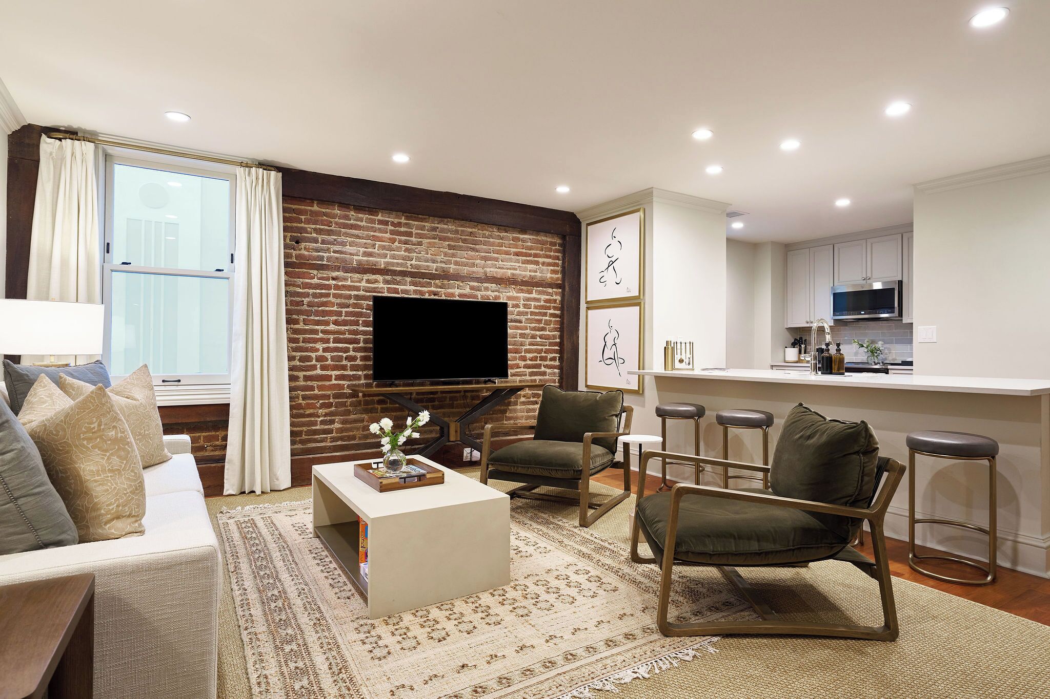 A modern living room with a brick wall, a flat-screen TV, a sofa, a coffee table, and a kitchen area with bar stools.