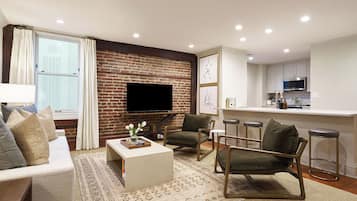 A modern living room with a brick wall, a flat-screen TV, a sofa, a coffee table, and a kitchen area with bar stools.