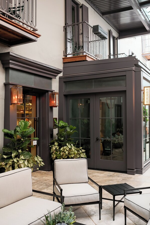 A modern hotel exterior with a glass door entrance, outdoor seating area with chairs, and potted plants.