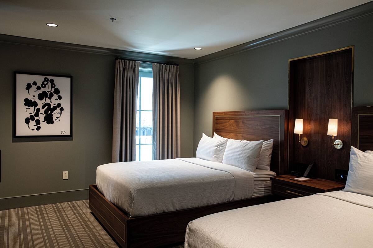 A hotel room with two beds, a large window with curtains, a framed artwork on the wall, and a wooden headboard with wall-mounted lights.