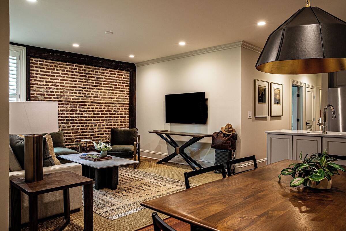 A modern living room with a brick wall, a flat-screen TV, a dining table, and a kitchen area with a stainless steel refrigerator.
