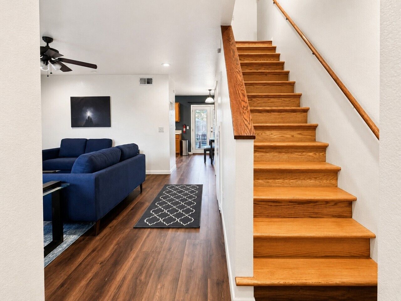 Una escalera con escalones de madera y barandilla, un sofá azul, una alfombra blanco y negro, y un ventilador de techo.