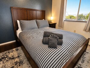A bed with striped bedding, a wooden headboard, a nightstand with a lamp, and a view through a window.
