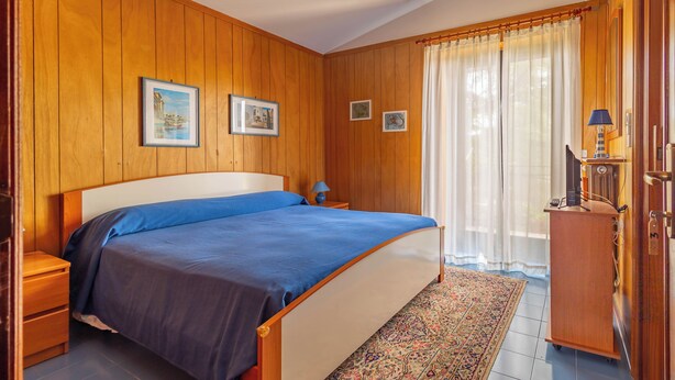 Un dormitorio con paneles de madera, una cama con ropa de cama azul, dos cuadros enmarcados en la pared, una mesita de noche, una lámpara y un radiador.