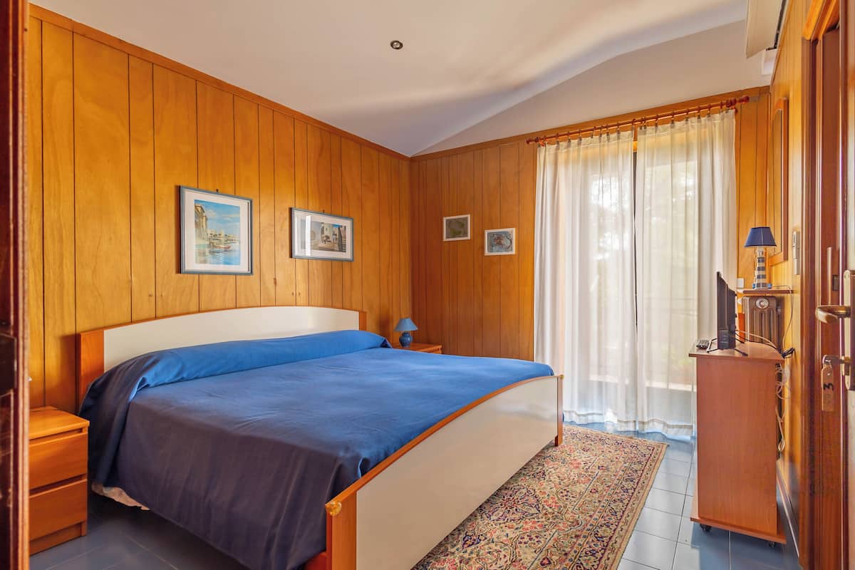 A bedroom with wooden paneling, a bed with blue bedding, two framed pictures on the wall, a nightstand, a lamp, and a radiator.