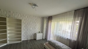 Een slaapkamer met een grote kledingkast, een nachtkastje met een lamp, een bed met bedkussens met een patroon, en een raam met dunne gordijnen.