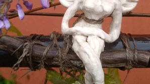 A white statue of a woman sitting on a branch with purple flowers.