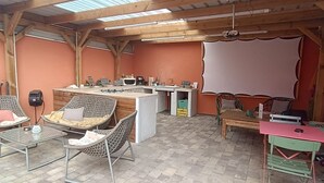 A rustic outdoor dining area with metal tables and wicker chairs, a kitchen counter, and a large white projection screen.