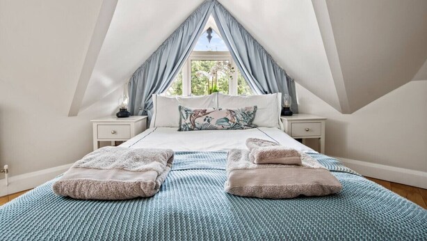 Un dormitorio con techo inclinado, una cama con colcha de diseño azul y blanco, dos almohadas y una mesita de noche con lámpara.