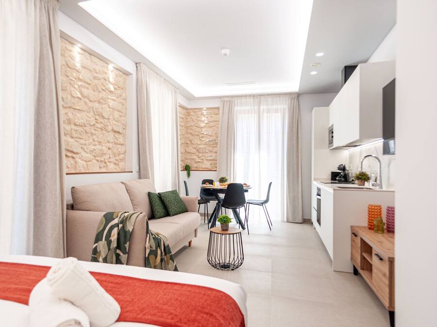 A modern hotel room with a bed, a sofa, a dining area with a table and chairs, and a kitchenette.