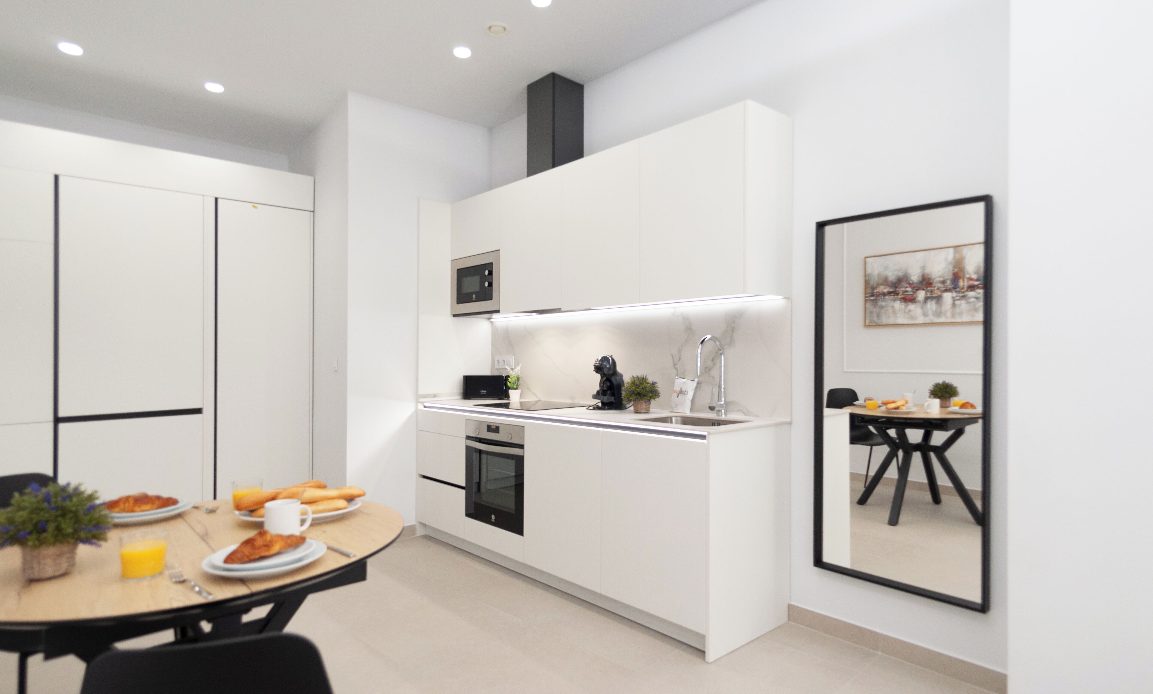 A modern kitchen with white cabinetry, a microwave, and a sink. There's a dining area with a table set for two, and a framed picture on the wall.