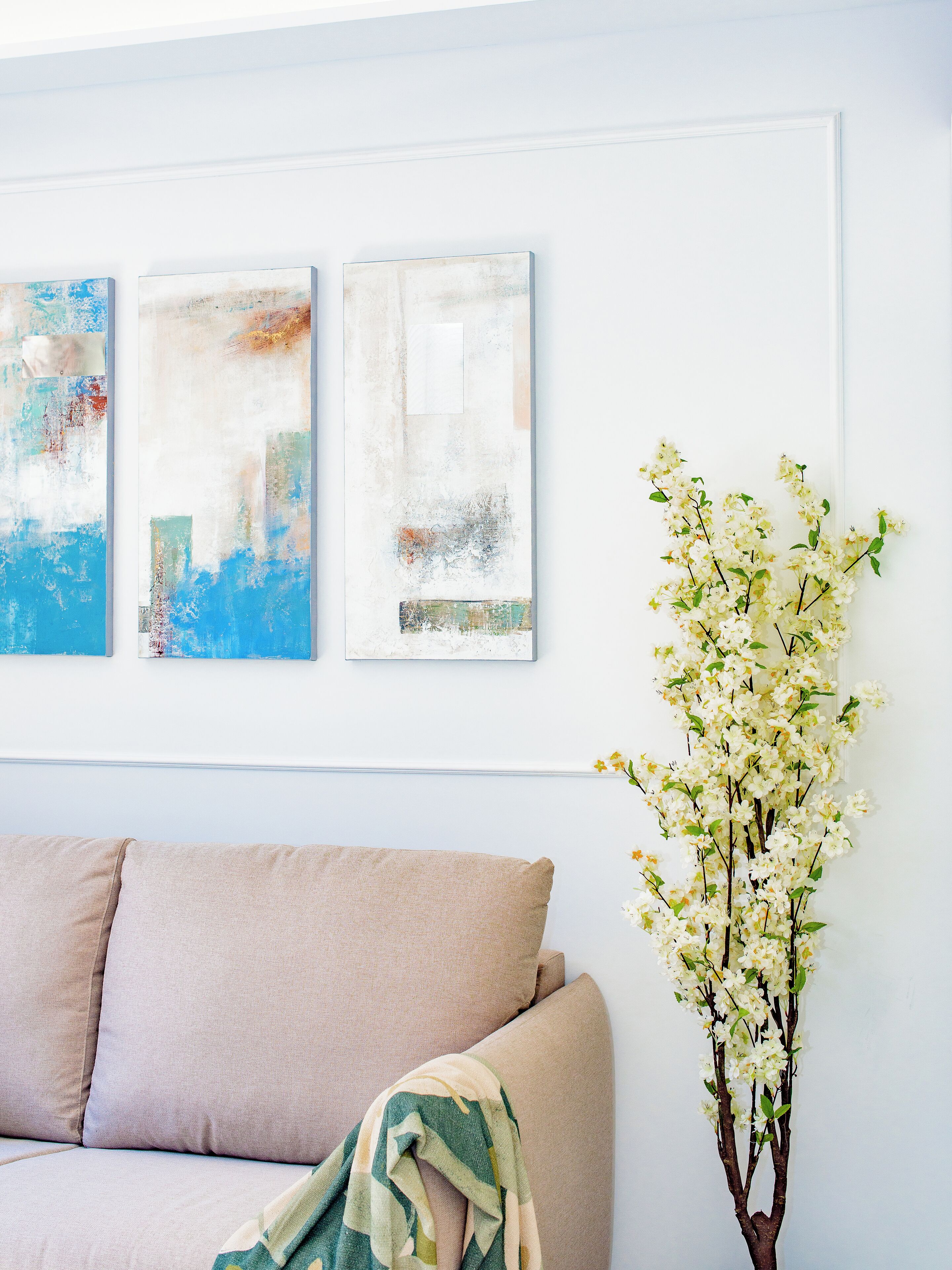 A living room with a beige sofa, three abstract paintings on the wall, and a vase with white flowers.