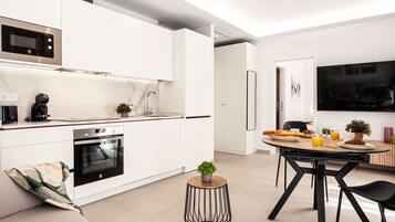 Een moderne keuken met witte kasten, een ingebouwde oven en een magnetron. Er is een eethoek met een tafel, stoelen en een klein rond bijzettafeltje met een plant.