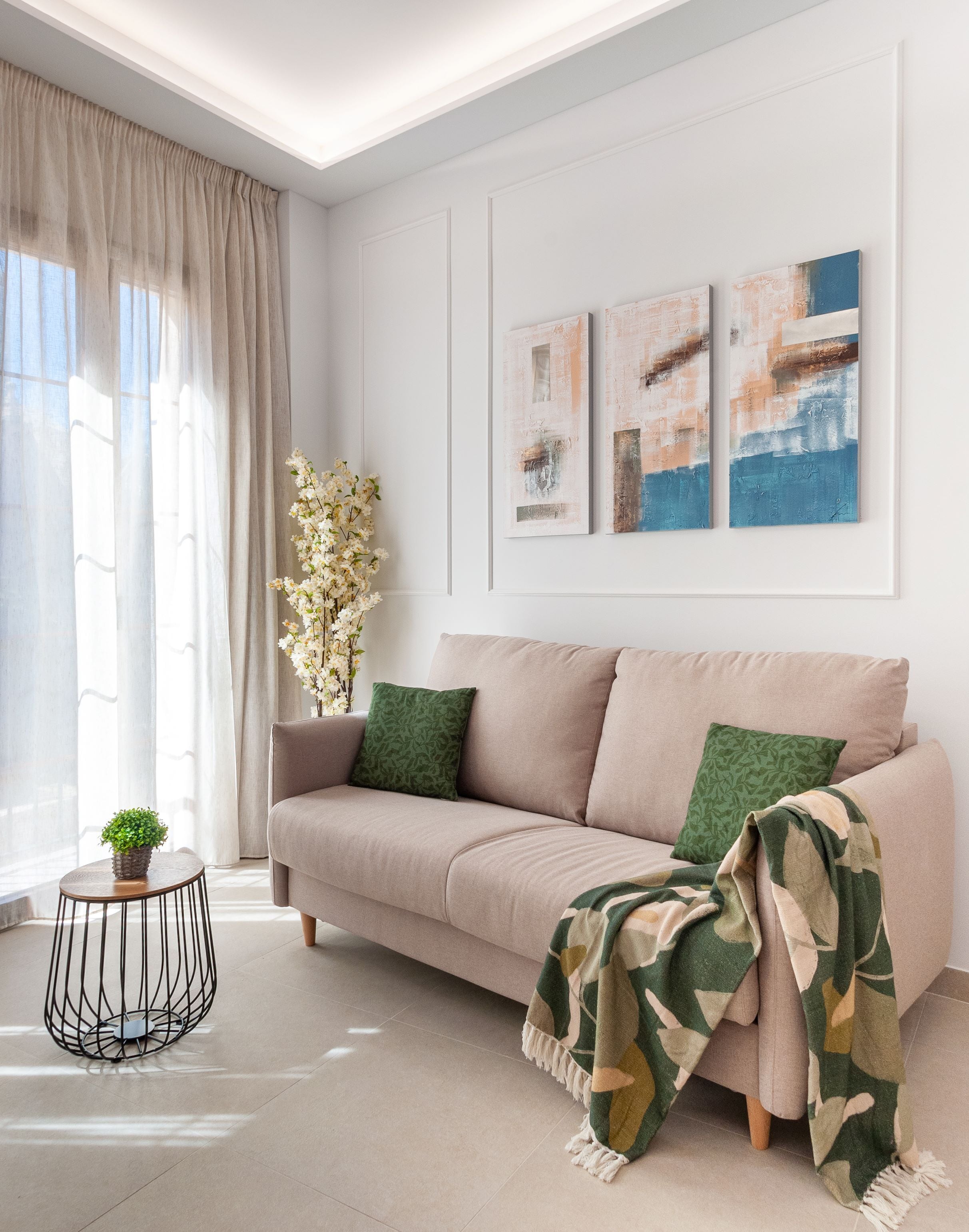 Un salón moderno con un sofá color beige, cojines verdes y una manta con estampado. Hay dos cuadros abstractos en la pared y una pequeña planta en maceta sobre una mesa redonda.