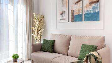 Un salón moderno con un sofá color beige, cojines verdes y una manta con estampado. Hay dos cuadros abstractos en la pared y una pequeña planta en maceta sobre una mesa redonda.