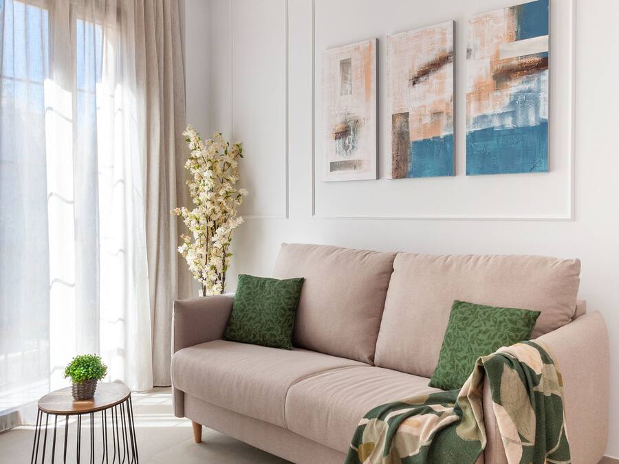 Un salón moderno con un sofá color beige, cojines verdes y una manta con estampado. Hay dos cuadros abstractos en la pared y una pequeña planta en maceta sobre una mesa redonda.