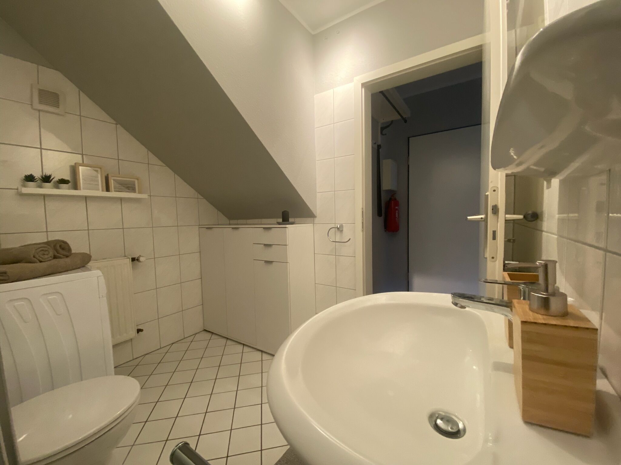 A bathroom with a white sink, toilet, and shower area. There are shelves with toiletries and a towel on the radiator.