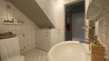 A bathroom with a white sink, toilet, and shower area. There are shelves with toiletries and a towel on the radiator.