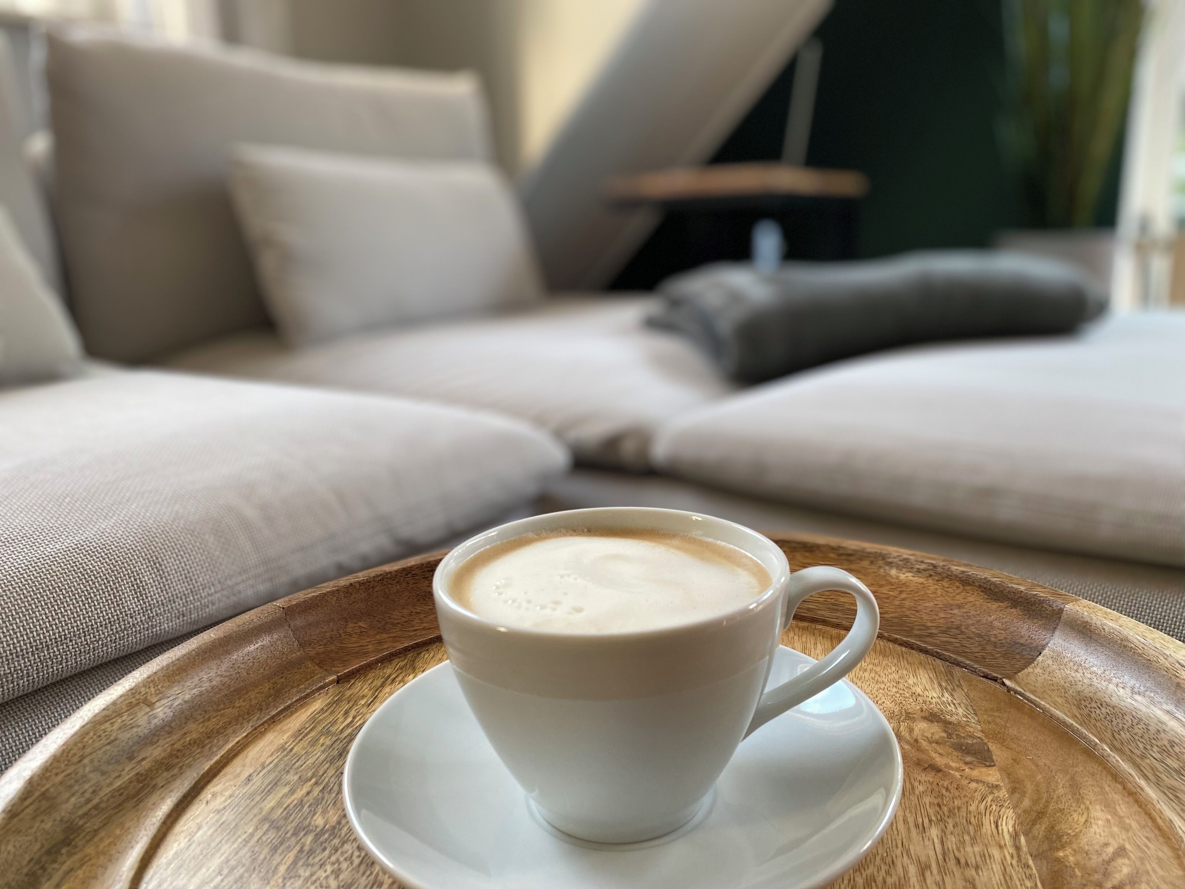 En kop kaffe på en træbakke foran en sofa med puder.