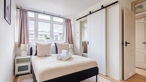 Un dormitorio con cama, mesitas de noche, una ventana con cortinas, una puerta corredera y vistas a edificios.