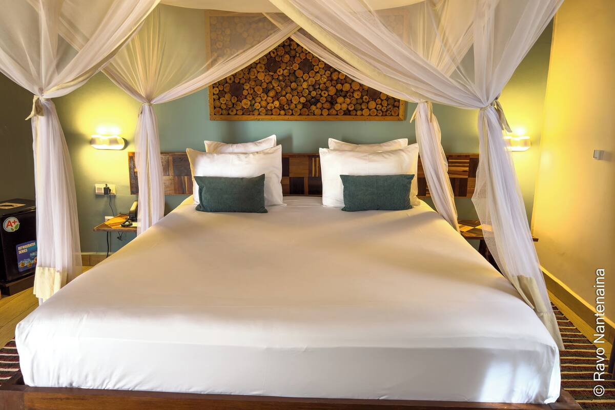 A large bed with white linens and two green pillows, a wooden headboard, and a wall-mounted log display.