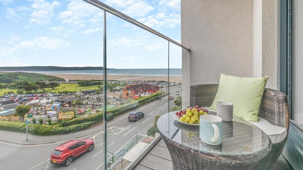 A balcony with a glass table, a wicker chair, and a view of a coastal town.