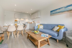 A modern living room with a blue sofa, wooden coffee table, and a dining area with chairs.