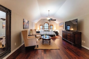 A spacious room with a large bed, a sitting area with a chair and a coffee table, a television, and a ceiling fan. - Lakeside Retreat | Fire Pit, Game Room & Canoe (Austin)