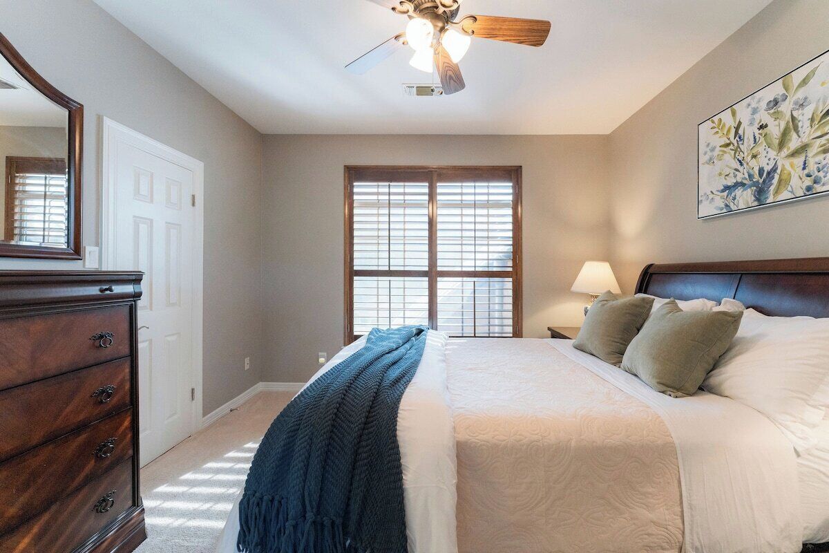 Une chambre à coucher comprenant un lit, une commode, un ventilateur de plafond, une fenêtre avec des stores et une lampe fixée au mur.