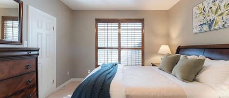 Une chambre à coucher comprenant un lit, une commode, un ventilateur de plafond, une fenêtre avec des stores et une lampe fixée au mur.
