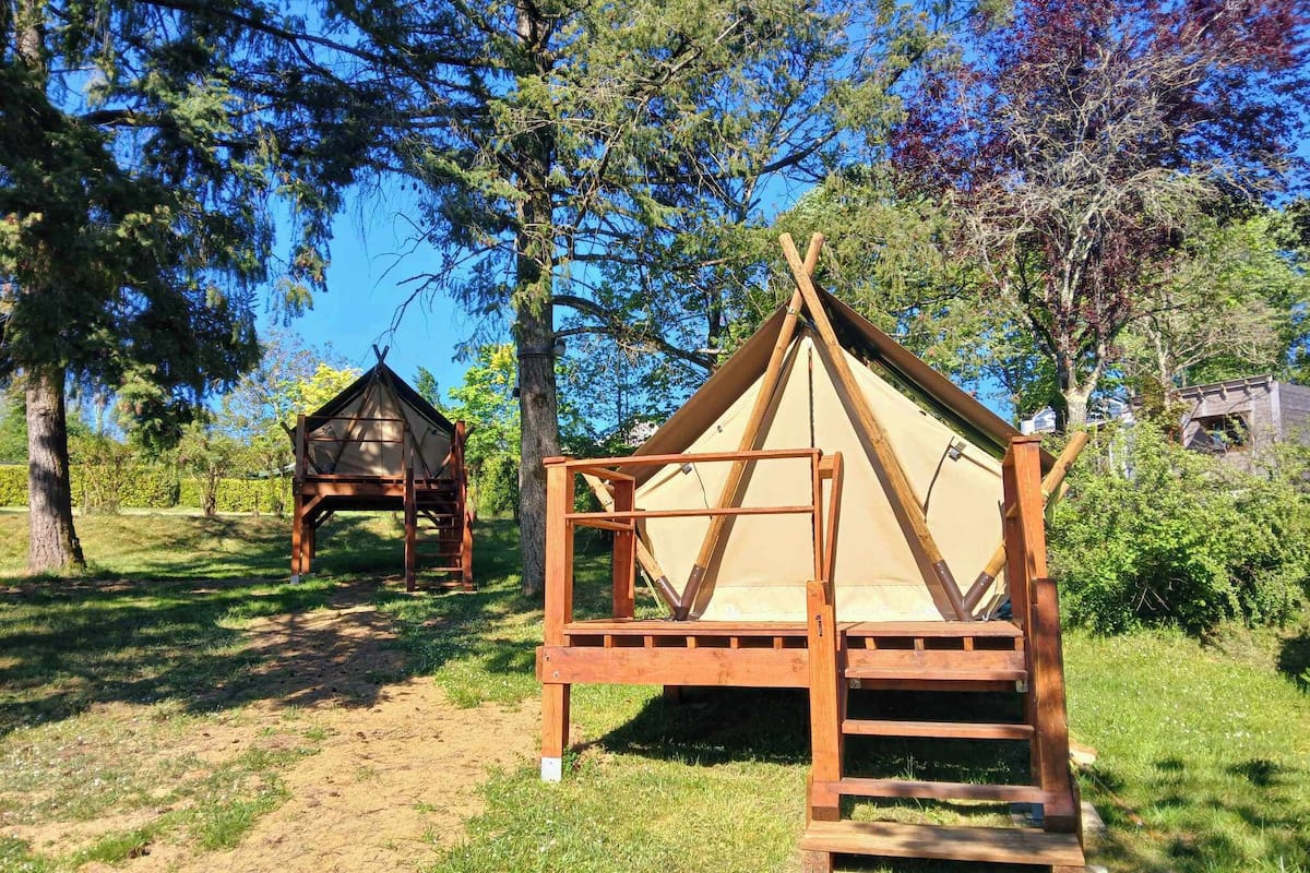 Two wooden tents with a simple design, set in a grassy area with trees in the background.