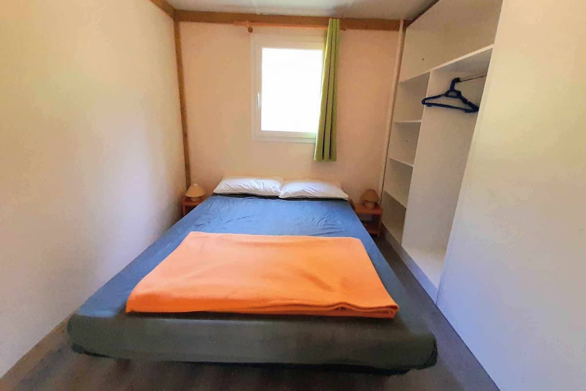 A small, narrow room with a single bed, a window with green curtains, and a small shelf with a hanger.