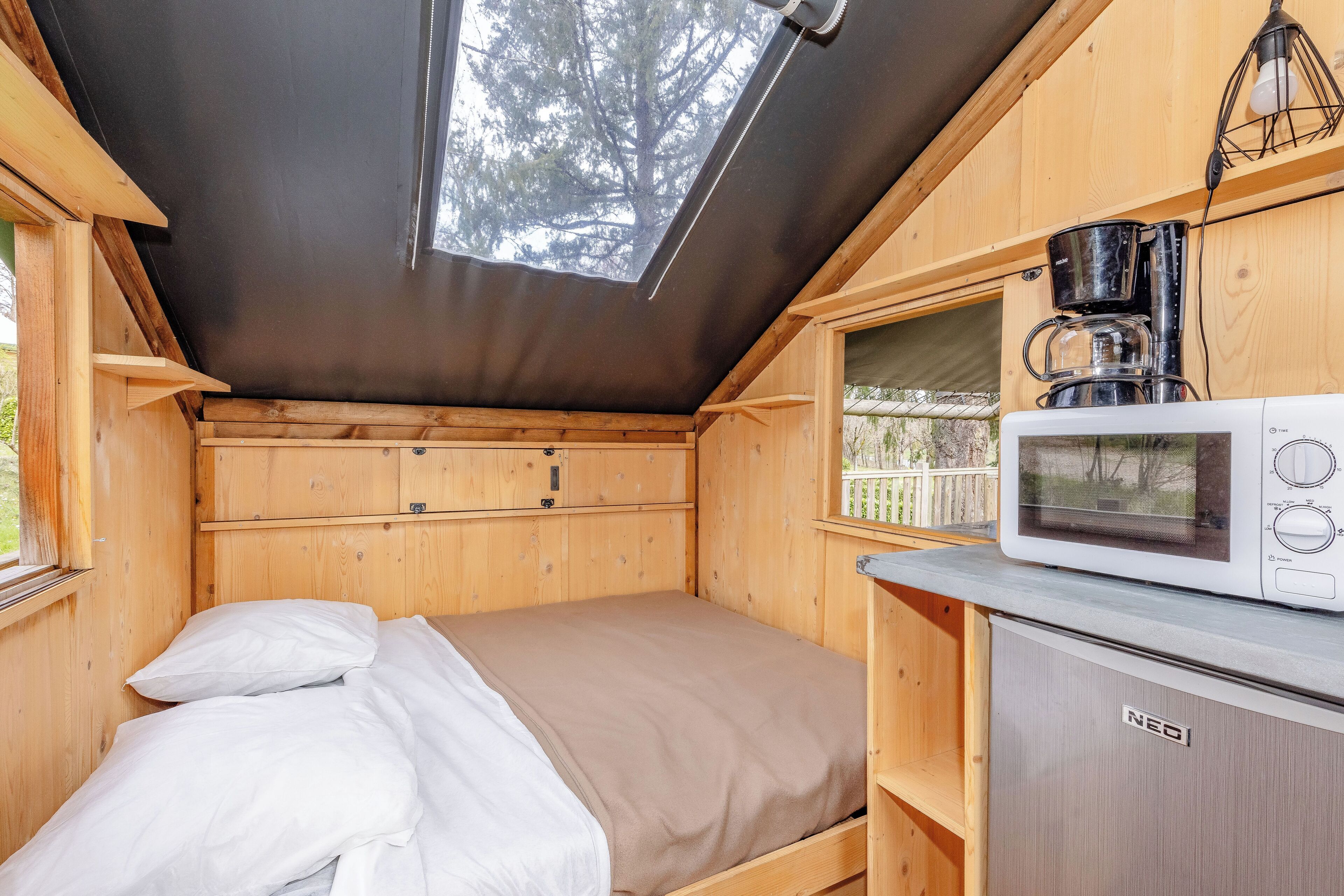 Una cabaña de madera pequeña con una cama, una kitchenette con microondas y una ventana con vista a un área cercada.