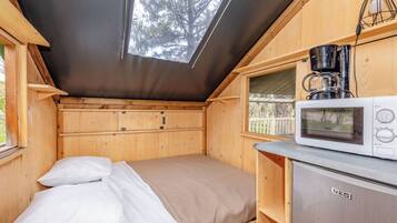 Una cabaña de madera pequeña con una cama, una kitchenette con microondas y una ventana con vista a un área cercada.