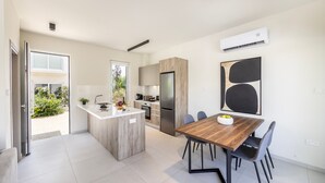 Una cocina moderna con una isla central, un comedor con mesa y sillas de madera, y un amplio ventanal que proporciona luz natural.