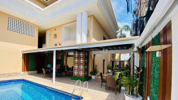 A swimming pool with a tiled deck, a covered seating area with chairs and tables, and potted plants. - Presken Oniru (Lagos)