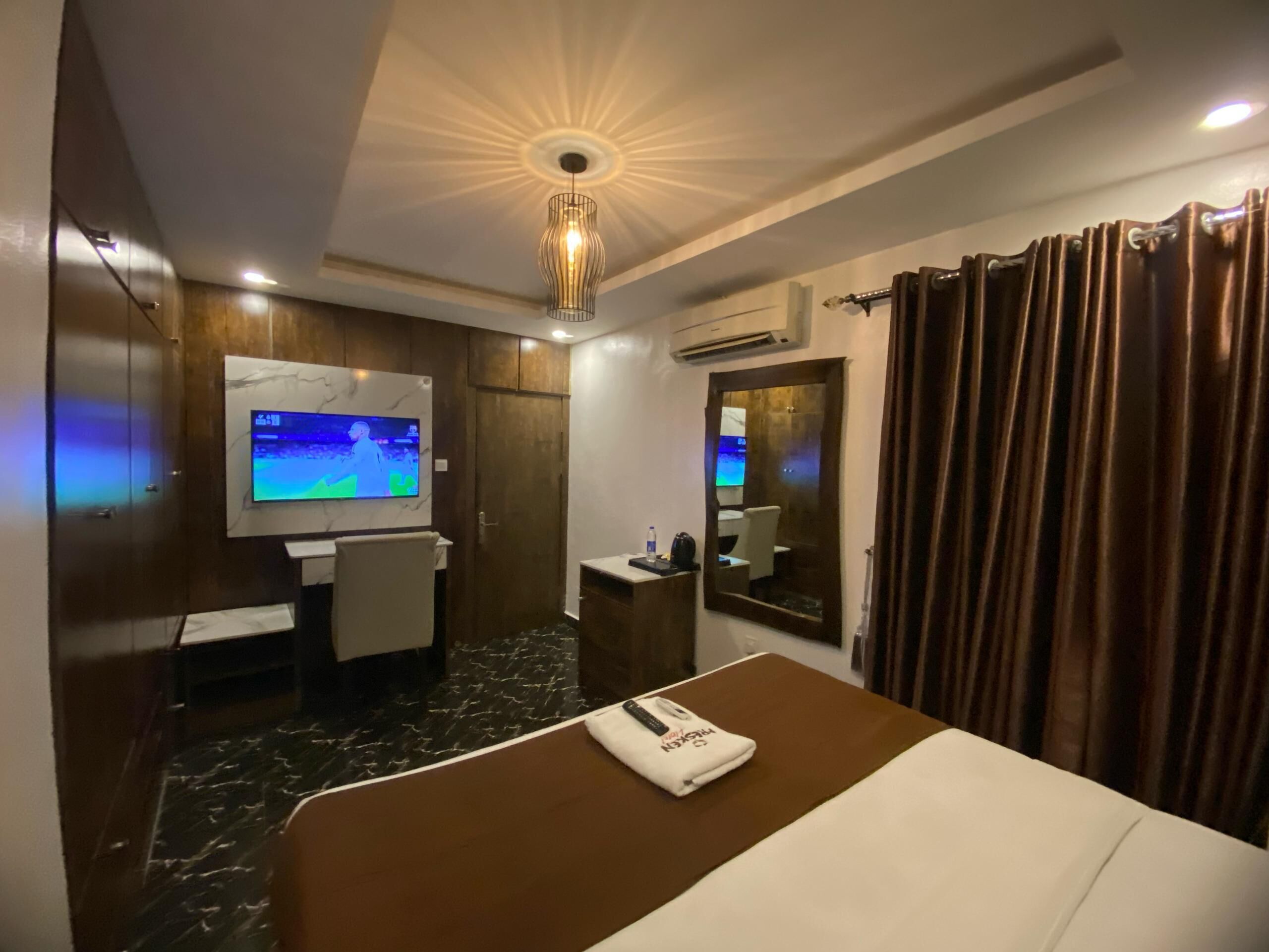 A hotel room with a bed, a TV mounted on the wall, a desk with a chair, a mirror, and a curtain.