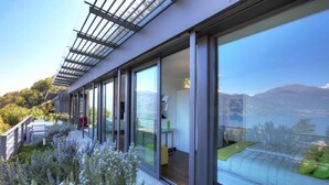 A modern building with large glass windows, a balcony with outdoor seating, and a view of a mountainous landscape. - FABULOUSLY LOCATED WITH VIEWS OF LAKE COMO WITH A HEATED POOL (Menaggio)
