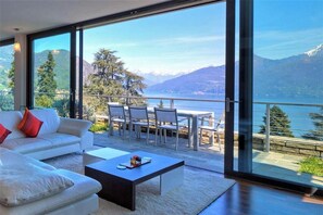 A modern living room with a large glass door leading to a balcony overlooking a lake and mountains. - FABULOUSLY LOCATED WITH VIEWS OF LAKE COMO WITH A HEATED POOL (Menaggio)