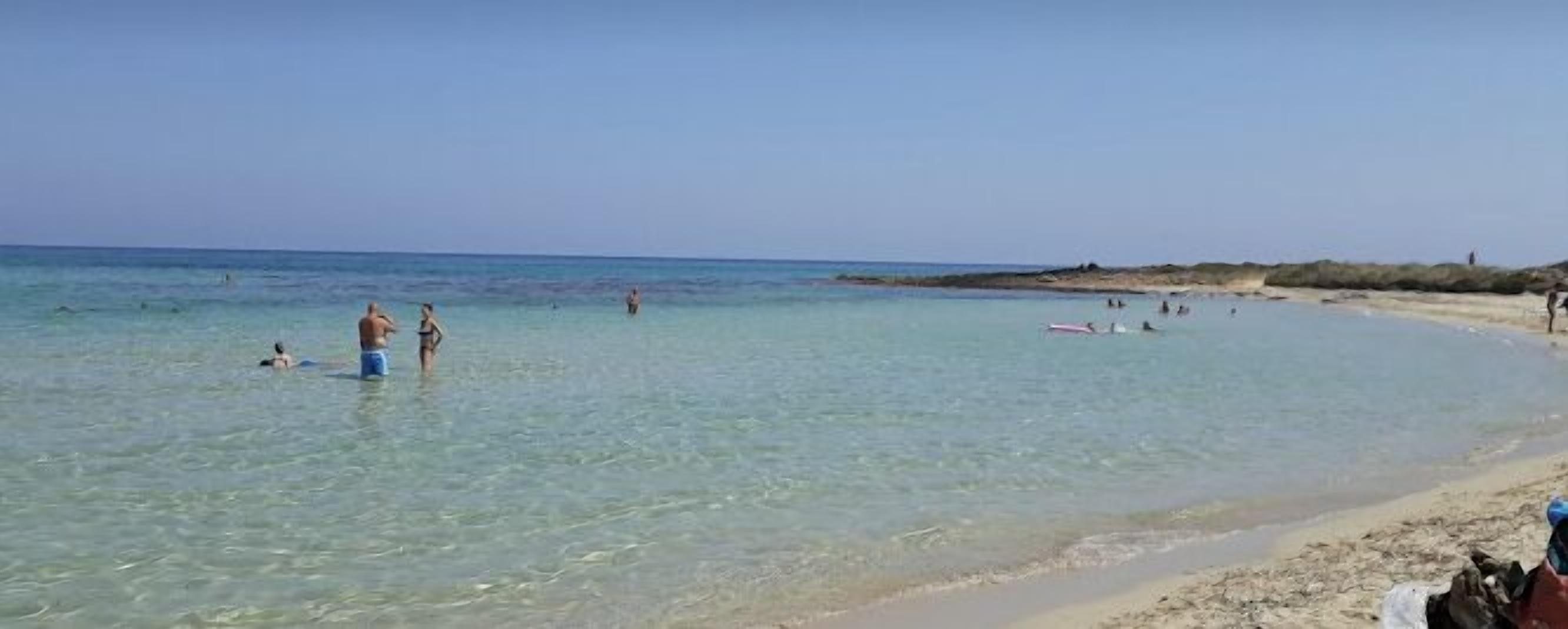 A beach with clear water, people swimming and sunbathing, and a sandy shore.